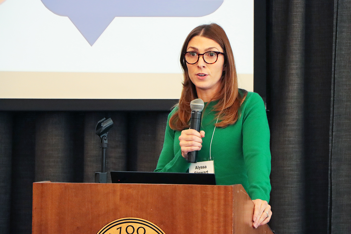 Alyssa Stewart at lectern in front of screen, with microphone and green shirt