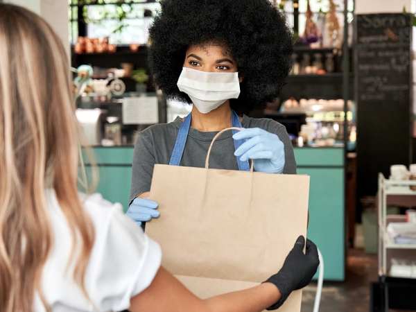 Black woman wearing surgical mask and gloves with beautiful 4C hair hands takeout bag to white woman customer in gloves
