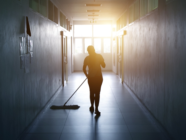 Custodian pushes a push broom in backlit hallway