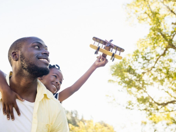 African-American father holds child flying toy airplane in bright sky