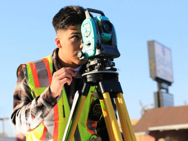 Young man surveyor in high-visibility vest looks through total station on tripod